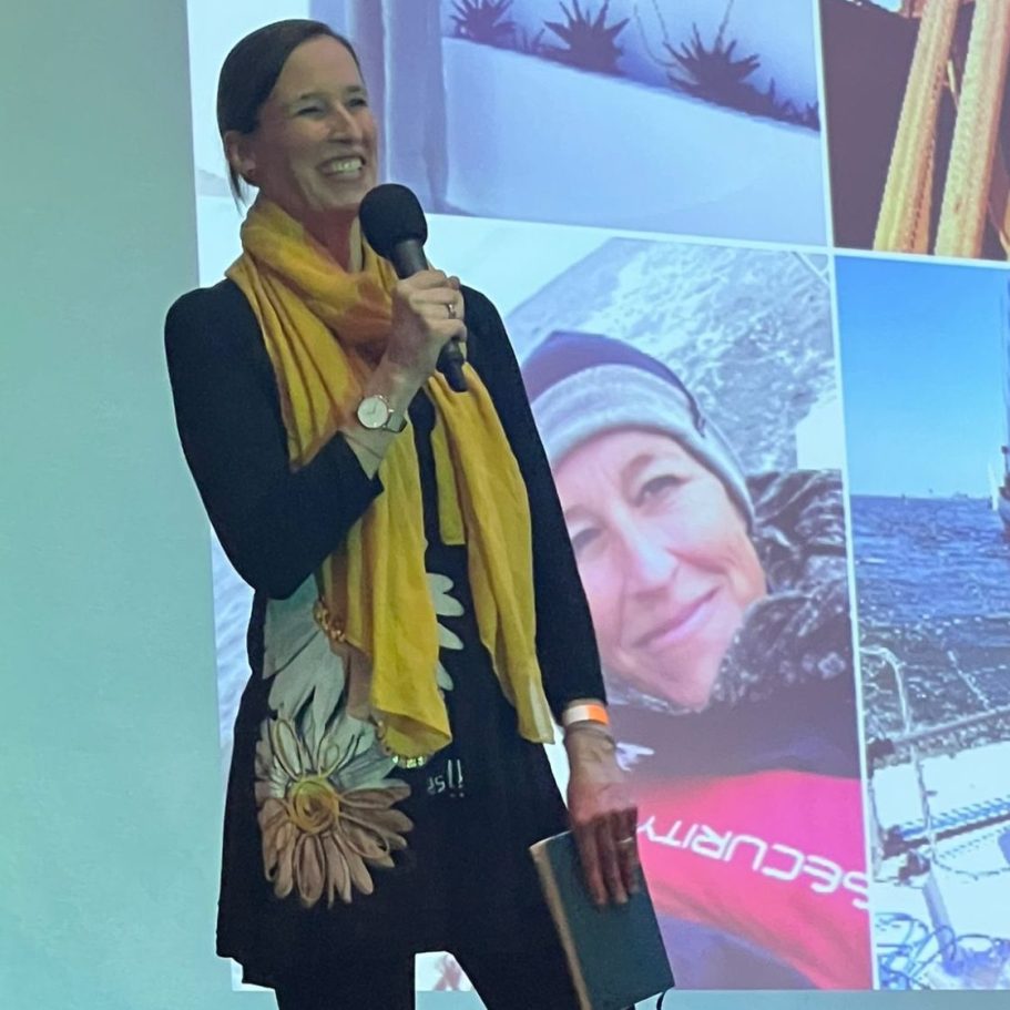 Präsenz Meike Völkner trägt ein geblümtes, schwarzes Kleid und hat ein Mikrophon in der Hand. Sie steht auf eine Bühne und lächelt.