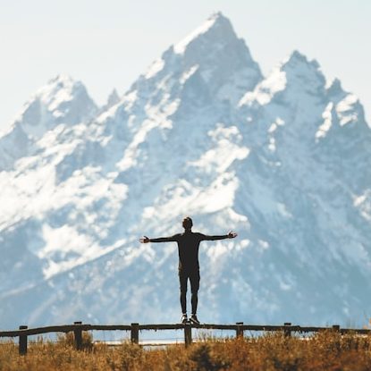 Mann mit ausgebreiteten Armen von hinten, der auf einem Zaun vor einem großen, schneebedeckten Berg balanciert 